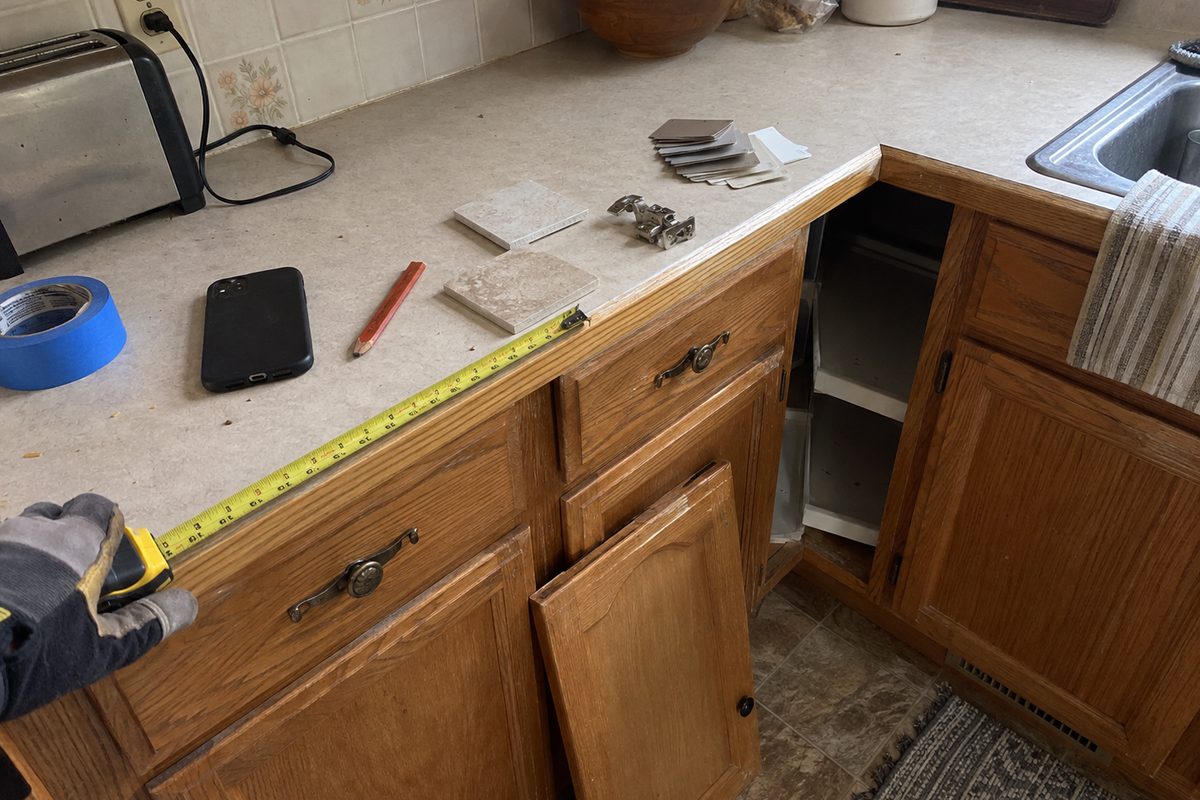 A gloved hand pulling a tape measure along an old oak kitchen cabinet, with a phone face-down, a carpenter pencil, tile samples, and a hinge on the laminate counter.