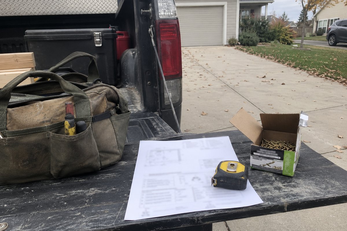 A one-page job sheet with its text blurred, held down by a tape measure on a pickup tailgate beside a toolbag and a box of screws.