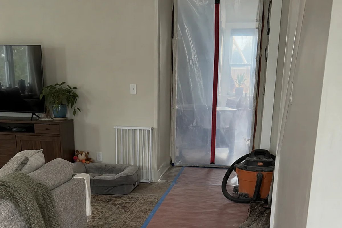 A living room with red rosin paper taped down over the floor leading to a plastic zip wall in the doorway, with a shop vacuum and dusty work boots beside it and a baby gate, dog bed and stuffed toy against the wall.