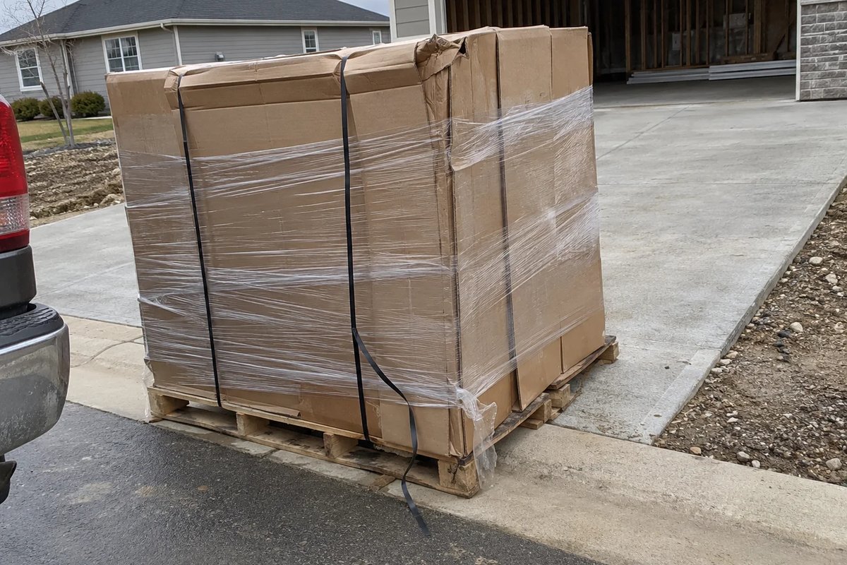 A shrink-wrapped pallet of cardboard-boxed materials dropped at the curb at the edge of a driveway, one box corner crushed and a black strap hanging loose, with a pickup bumper at the left edge and an open garage behind.