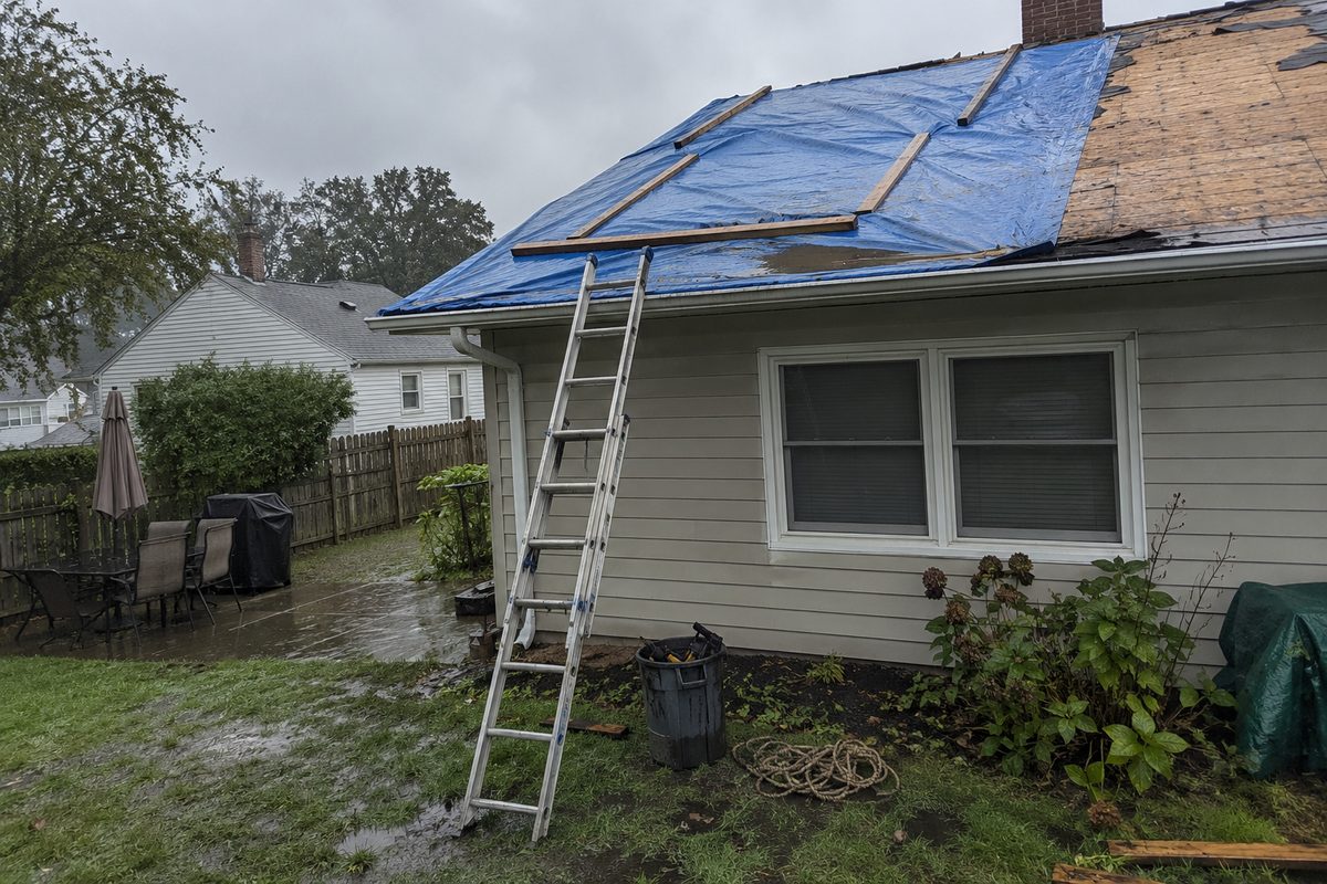 A half-stripped house roof covered with a wet blue tarp held down by boards, a ladder against the gutter, and a bucket of tools on a soggy lawn under gray skies.