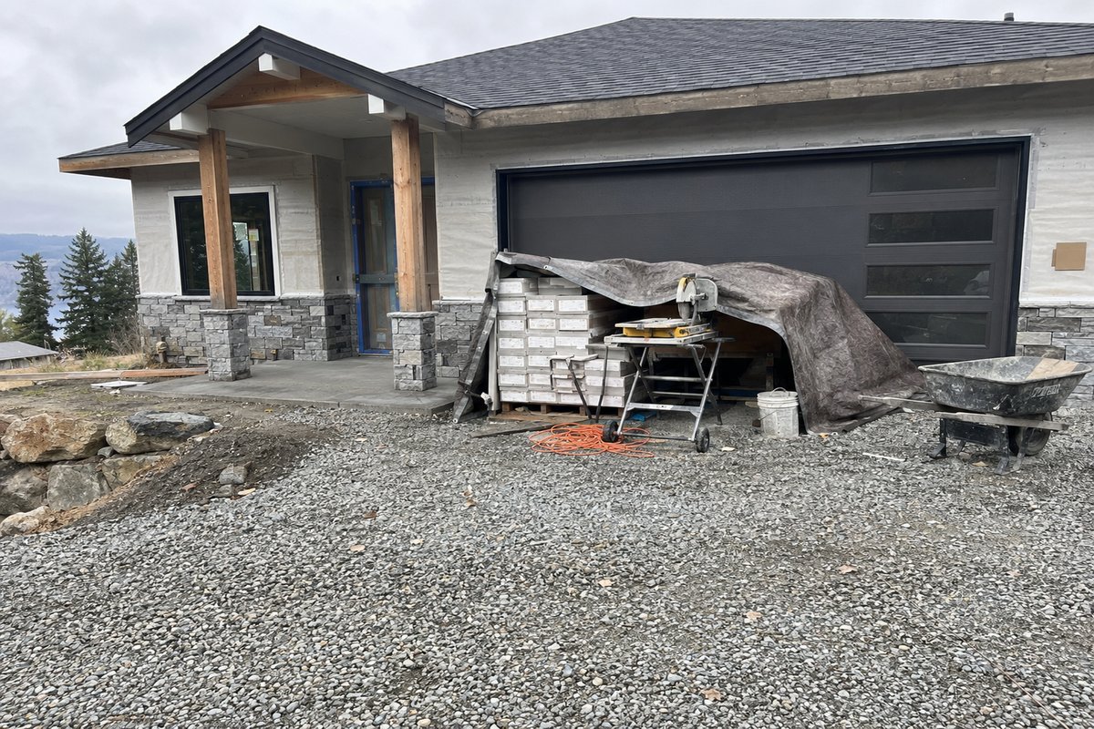 A new house with an empty gravel driveway, a tarp-covered pallet of tile boxes and a wet saw on a stand by the garage door, an orange extension cord and a wheelbarrow, and no vehicle or worker in sight.