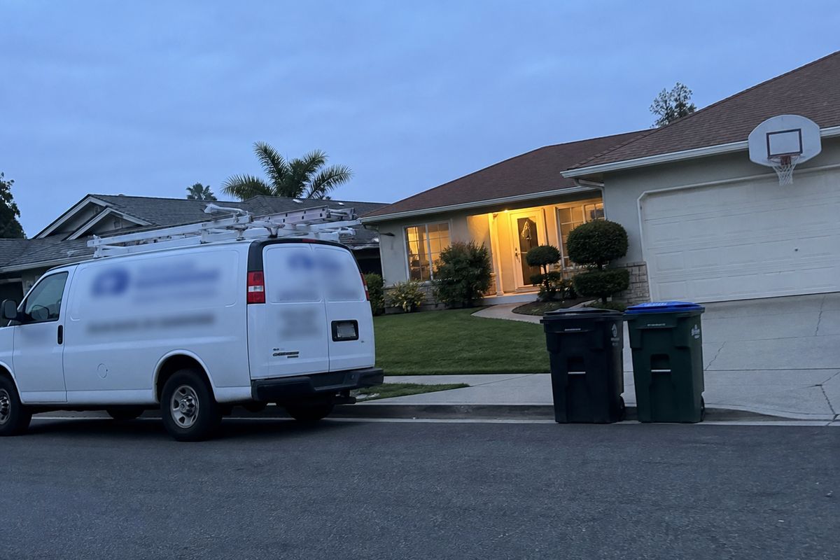 A white work van parked at the kerb outside a suburban house at dusk with the porch light on.